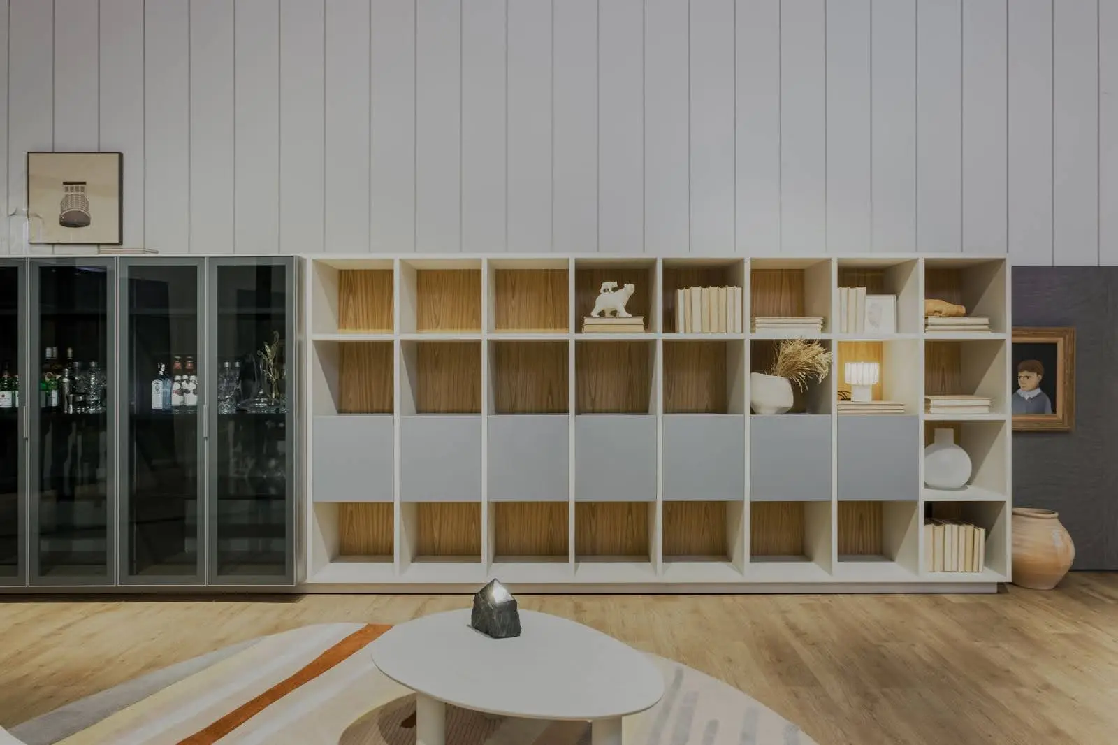 Estante moderna Evivva com nichos abertos e fechados exibindo livros, vasos e objetos decorativos. Armário com portas de vidro mostra bebidas e taças; mesa de centro branca.